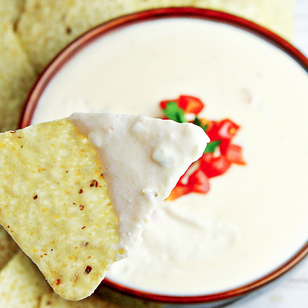Tortilla chip being dipped into Homemade White Queso Dip in a brown bowl, and topped with diced tomatoes