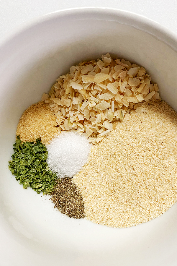 A white bowl with six seasonings in the bottom: onion powder, onion flakes, garlic powder, salt, pepper, and parsley