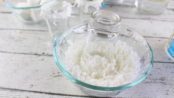 Glass shot glass that has vanilla frosting on the rim being dipped into Shredded Sweetened Coconut