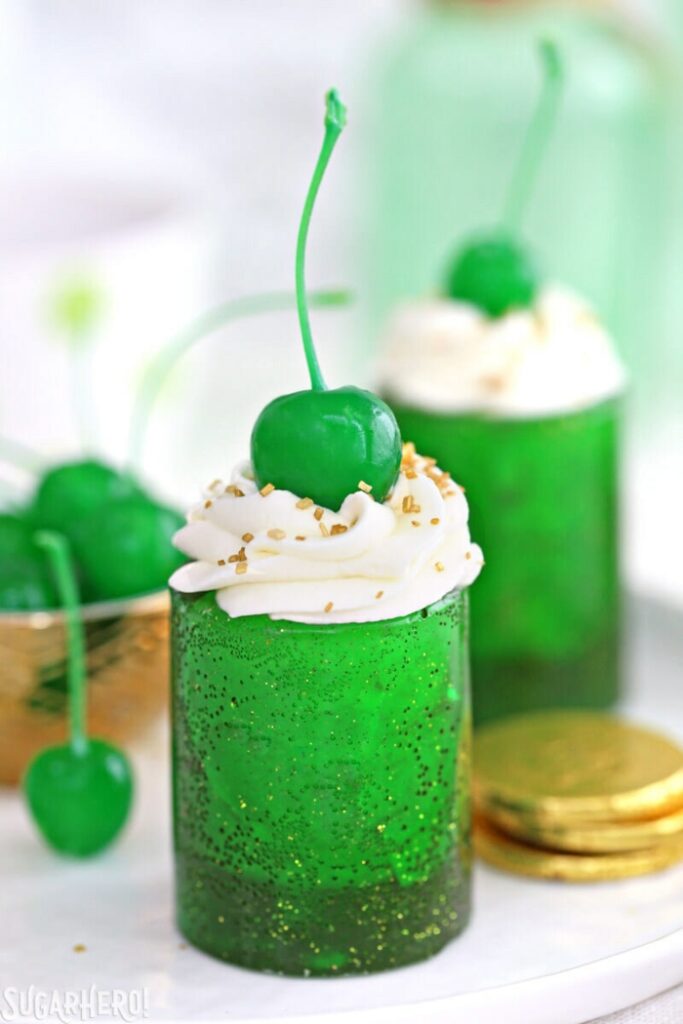 Two Shamrock Shake Shots on a white surface with a bowl of green cherries next to the shots.