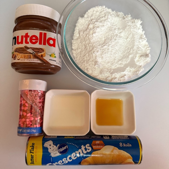 The ingredients to make Valentine's Day Nutella Crescents are Nutella, Crescents rolls, powered sugar, Valentine’s Day sprinkles, milk and vanilla extract.
